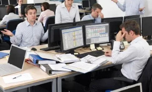 Open-plan office with coworkers; a man presents at a whiteboard while others work at desks.