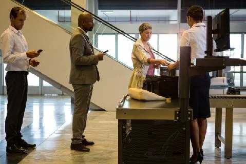 Receptionist checks a visitor's documents at a desk while four businesspeople wait in a modern office lobby.