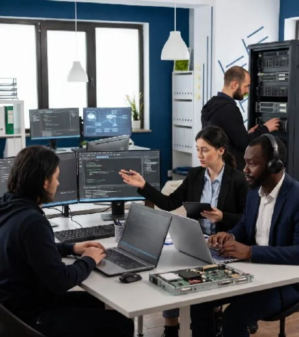 Team of four IT professionals collaborating in a modern lab: two coding at laptops, one presenting with a tablet, a server rack in the background.