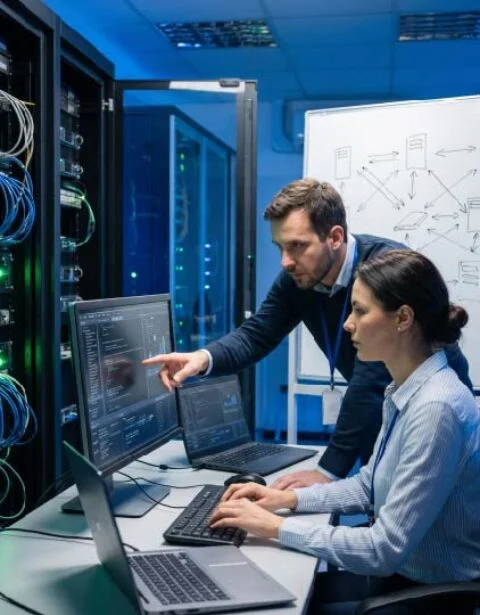 Two IT professionals work at a server rack, one points at a monitor while the other types at a laptop.