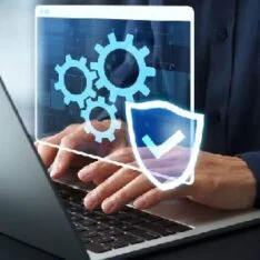 Person typing on a laptop with blue gear icons and a shield with a checkmark, symbolizing cybersecurity.