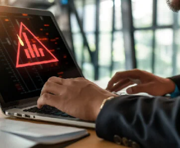 Person in a suit typing on a laptop that shows a red warning triangle and data graphs, signaling a cybersecurity alert.